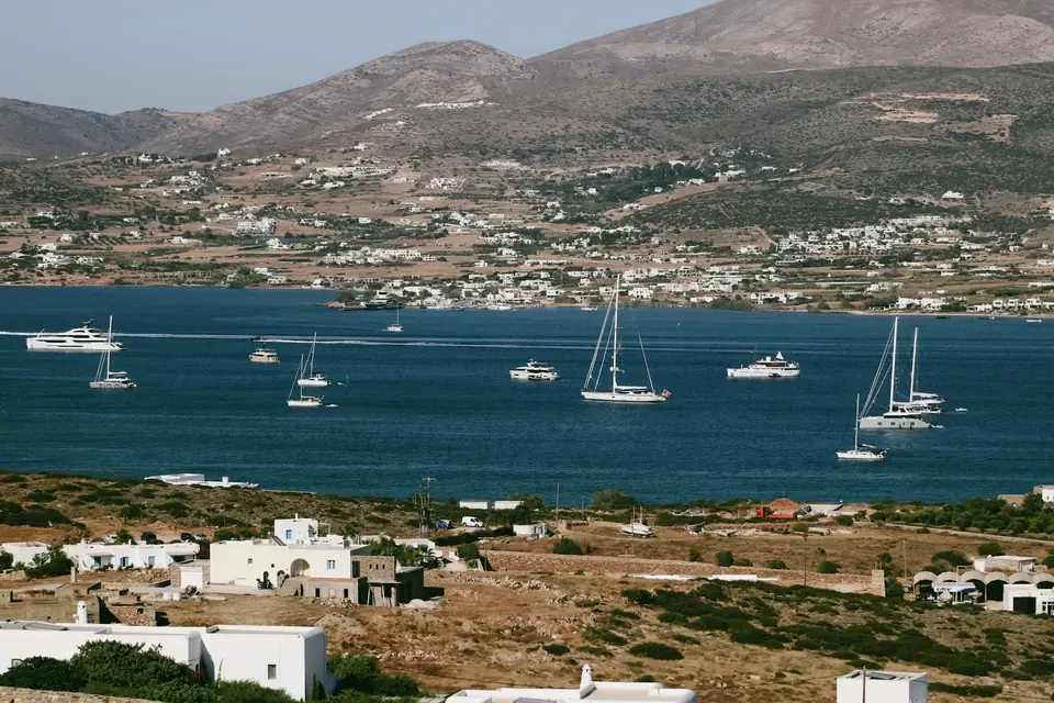 Sailboats and yachts across a blue bay, with white houses and dry hills beyond.